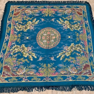 Puede incluir: Un tejido cuadrado de color verde azulado con un medallón circular central de motivos florales, rodeado de grullas bordadas, pinos y formaciones rocosas. El borde está decorado con patrones adicionales de grullas y flores, con flecos.