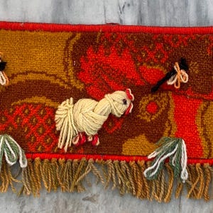 May include: A rectangular, vintage-style wall hanging with a repeating pattern of stylized chickens and floral designs. The piece features a warm color palette of orange, brown, and yellow, with a fringed bottom edge. The chickens are made of cream-colored yarn.