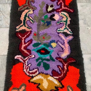 May include: A rectangular, abstract rug with a dark gray border and a central design in shades of purple, orange, and green. The rug features a shaggy texture and a unique, artistic pattern, suitable for home decor.