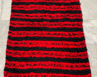 May include: A rectangular rug with alternating horizontal stripes of red and black. The rug has a shaggy texture and is made of a soft material. The top edge has several wooden toggles for hanging.