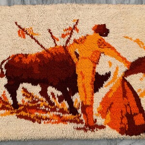 May include: A beige rug with a vibrant depiction of a bullfight scene. The image features a bull, a matador, and spears, rendered in shades of brown, orange, and yellow. The rug has a textured, shaggy appearance.
