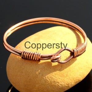 Può includere: Un braccialetto a cerchio color rame con chiusura a gancio. Il braccialetto è realizzato con filo avvolto e presenta la scritta "Coppersty". Il braccialetto è esposto su una pietra marrone chiaro.