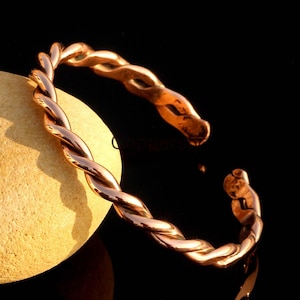 Könnte beinhalten: Ein kupferfarbenes, gedrehtes Metallarmband. Das offene Armband hat ein geflochtenes Design mit poliertem Finish. Das Armband wird auf einem hellen, abgerundeten Stein vor schwarzem Hintergrund präsentiert.