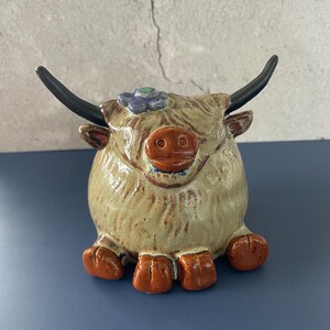 May include: A ceramic figurine of a Highland cow with a round, beige, textured body, dark brown horns, and a brown snout. The cow features a small purple flower detail on its head and four brown hooves at the base, creating a compact, seated animal sculpture.