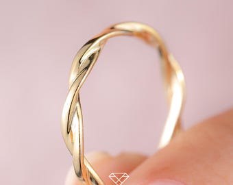 Puede incluir: Un anillo dorado con un diseño de cuerda trenzada. El anillo está sostenido por los dedos de una persona sobre un fondo rosa suave. El diseño del anillo presenta hebras entrelazadas, creando una apariencia texturizada. El anillo es una joya.