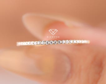 Puede incluir: Un anillo de banda fina de oro con una hilera continua de pequeñas piedras claras de corte redondo engastadas en la parte superior. El anillo muestra un pequeño logotipo con el texto DIAMOND CORA centrado sobre la banda. Acabado pulido.