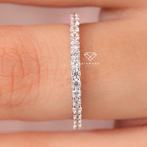 Puede incluir: Primer plano de un anillo de diamantes de la eternidad. El anillo está engastado con pequeños diamantes redondos y transparentes. El anillo está en un dedo, con el tono de la piel en el fondo. Es una joya delicada.