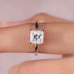 以下が含まれることがあります： 四角いカットのダイヤモンドが付いたシルバーの婚約指輪。 ダイヤモンドは4本爪でセットされ、バンドは細くて磨かれています。 リングは指にあり、背景はソフトピンクです。