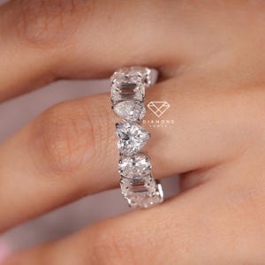 May include: A close-up of a diamond eternity ring featuring a mix of emerald, pear, and heart-shaped stones. The ring is set in a white metal, and the stones sparkle brightly. The ring is on a finger.