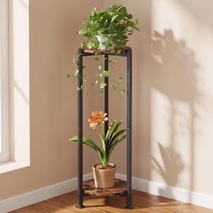 May include: A tall, corner-shaped plant stand featuring a black metal frame with three vertical legs and two circular, dark wood-grain shelves. A green leafy plant sits on the top shelf, and a potted orange flower sits on the bottom shelf.