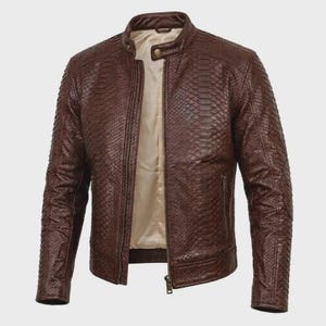 Könnte beinhalten: Eine braune Jacke mit einer strukturierten Schlangenledermusterung auf der Außenseite. Die Jacke verfügt über einen Stehkragen mit einem Druckknopfverschluss, einen durchgehenden Frontreißverschluss und zwei Seitentaschen mit Reißverschlüssen.