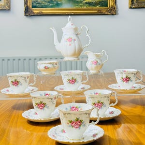 Puede incluir: Un juego de té vintage con un diseño de rosas. El juego incluye una tetera, una jarra de leche, un azucarero y varias tazas de té con platillos. El juego de té se exhibe sobre una mesa de madera, con pinturas de paisajes enmarcadas en el fondo.