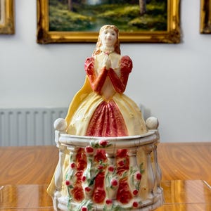 May include: A vintage porcelain figurine of a woman in a yellow and red dress, hands clasped in prayer. The figurine sits atop a decorative base with red rose accents. The background features framed landscape paintings.