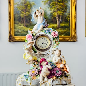 Puede incluir: Un reloj de porcelana blanca con figuras de querubines y adornos florales. La esfera del reloj es redonda con números y manecillas negras. La base y el marco están adornados con flores coloridas y ribetes dorados. El reloj está colocado frente a una pintura de paisaje enmarcada.