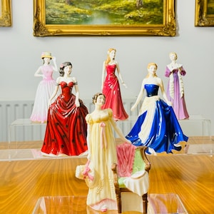 Puede incluir: Una colección de figuras de porcelana que representan a mujeres con elegantes vestidos. Las figuras son de varios colores, incluyendo rojo, rosa, azul y amarillo. El fondo presenta pinturas de paisajes enmarcadas con marcos dorados.