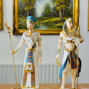 Op de afbeelding: Twee porseleinen Egyptische beeldjes. Eén figuur draagt een blauwe hoofdtooi en houdt een staf met een vogel erop. De andere figuur draagt een gouden en blauwe hoofdtooi en houdt een staf en een ankh vast. Beide figuren dragen witte gewaden met gouden en blauwe accenten.