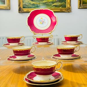 Pode incluir: Um conjunto de chá vintage com seis xícaras e pires, além de um prato de servir. O conjunto apresenta um esquema de cores vermelho escuro e dourado com um design floral no centro do prato e das xícaras. As xícaras e pires são exibidos em um suporte de acrílico transparente.