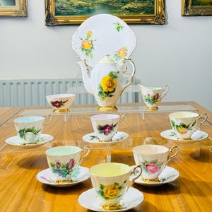 Peut inclure: Un ensemble de service à thé vintage comprenant une théière blanche, un pot à lait, un sucrier, et plusieurs tasses et soucoupes. L'ensemble est décoré de roses jaunes et de bordures dorées. Une assiette de service blanche avec des motifs de roses est également incluse.