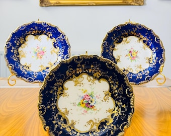 May include: Three antique porcelain plates with a dark blue border and gold filigree detailing. Each plate features a central floral bouquet in shades of pink, yellow, and blue. The plates are displayed on gold stands, with framed landscape paintings in the background.