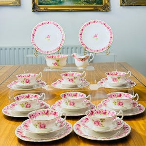 Puede incluir: Un juego de té vintage con un diseño de rosas rosas. El juego incluye tazas de té, platillos, platos, una jarra de crema y un azucarero. El juego está dispuesto sobre una mesa de madera. Se ven pinturas de paisajes enmarcadas en el fondo.