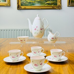 Puede incluir: Un juego de té vintage con un diseño floral. El juego incluye una tetera, una crema, un azucarero y cuatro tazas de té con platillos. Las tazas y los platillos están colocados sobre una mesa de madera. El juego es blanco con detalles florales rosas y morados.