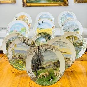 May include: A collection of decorative plates with landscape scenes, displayed on gold-colored stands. Each plate features a different pastoral image with a gold rim. The plates are arranged in a group, showcasing various nature-themed designs.