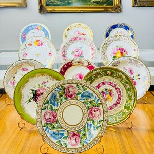 Op de afbeelding: Een verzameling antieke decoratieve borden met bloemmotieven. De borden hebben verschillende kleuren, waaronder roze, blauw, groen en goud. Elk bord wordt gepresenteerd op een goudkleurige standaard, die de ingewikkelde details en patronen benadrukt.