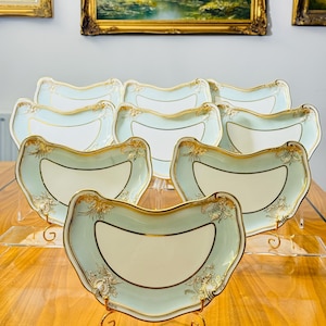 May include: A set of antique porcelain serving dishes. Each dish has a light blue and cream colour scheme with gold trim and floral accents. The dishes are displayed on clear stands, with landscape paintings in the background.
