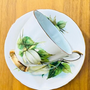 May include: White teacup and saucer set with a floral design. The cup and saucer are white with a light blue interior and gold trim. The floral design features white flowers and green leaves.