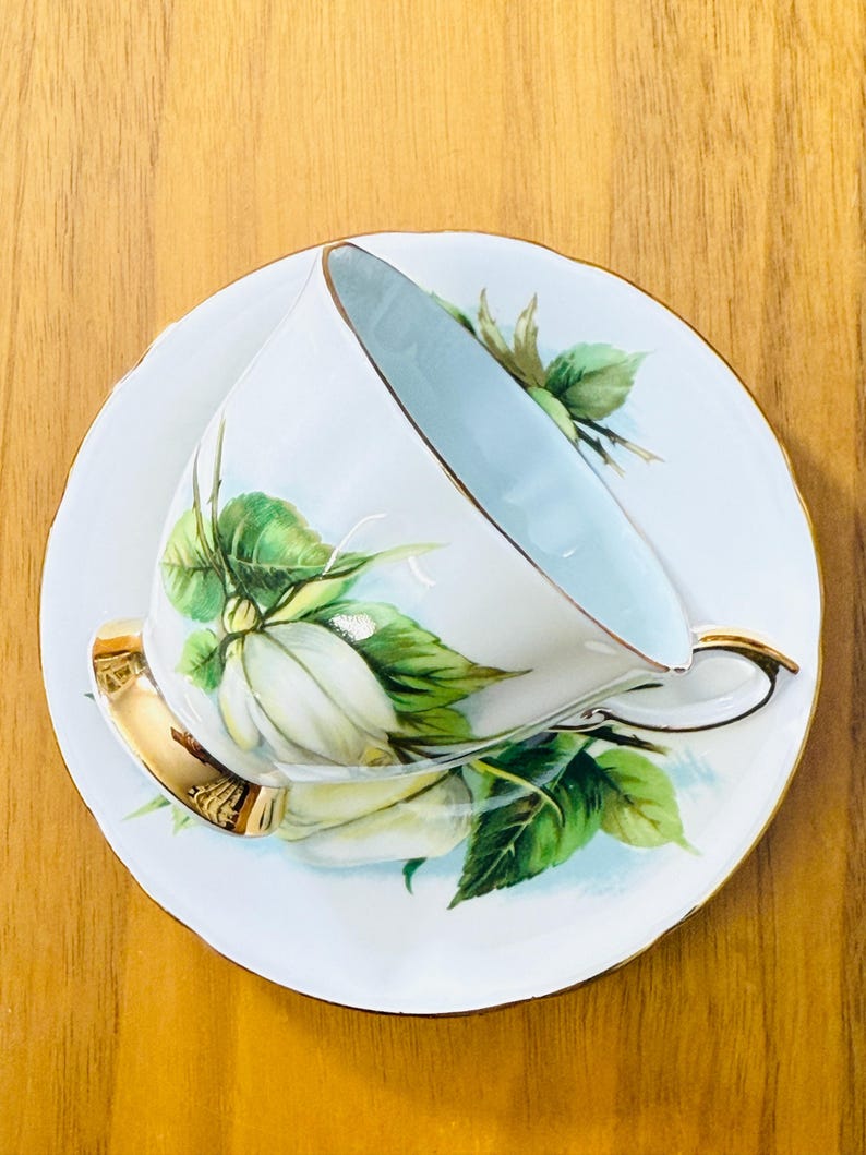 May include: White teacup and saucer set with a floral design. The cup and saucer are white with a light blue interior and gold trim. The floral design features white flowers and green leaves.