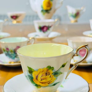 May include: A collection of vintage teacups and saucers with a floral rose design. The teacups have a yellow interior and gold trim. A teapot and creamer are also visible in the background. The teacups are on a wooden surface.