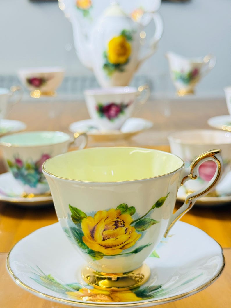 May include: A collection of vintage teacups and saucers with a floral rose design. The teacups have a yellow interior and gold trim. A teapot and creamer are also visible in the background. The teacups are on a wooden surface.