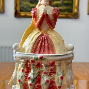 May include: A porcelain figurine of a woman in a long dress, with a yellow cape and red sleeves. The figurine is standing on a balcony decorated with red roses and green vines. The woman is holding her hands together in prayer.