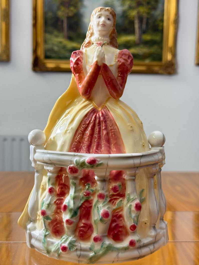 May include: A porcelain figurine of a woman in a long dress, with a yellow cape and red sleeves. The figurine is standing on a balcony decorated with red roses and green vines. The woman is holding her hands together in prayer.