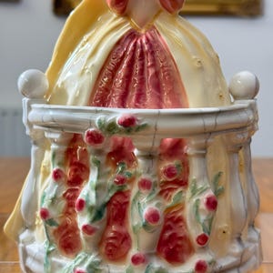 May include: A porcelain figurine featuring a woman in a yellow and red dress, standing behind a white balustrade decorated with red roses and green leaves. The figurine has a glossy finish and is set against a blurred background.