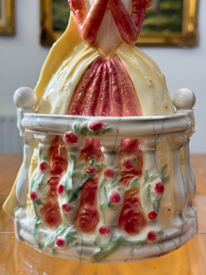 May include: A porcelain figurine featuring a woman in a yellow and red dress, standing behind a white balustrade decorated with red roses and green leaves. The figurine has a glossy finish and is set against a blurred background.