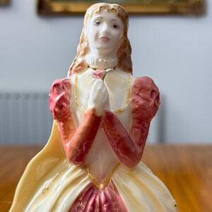 May include: A porcelain figurine of a woman in a long gown, with a cream-colored bodice and a pink skirt. The sleeves are a deep red, and she holds a pink rose. The figurine has long, flowing, light brown hair and is adorned with gold accents.