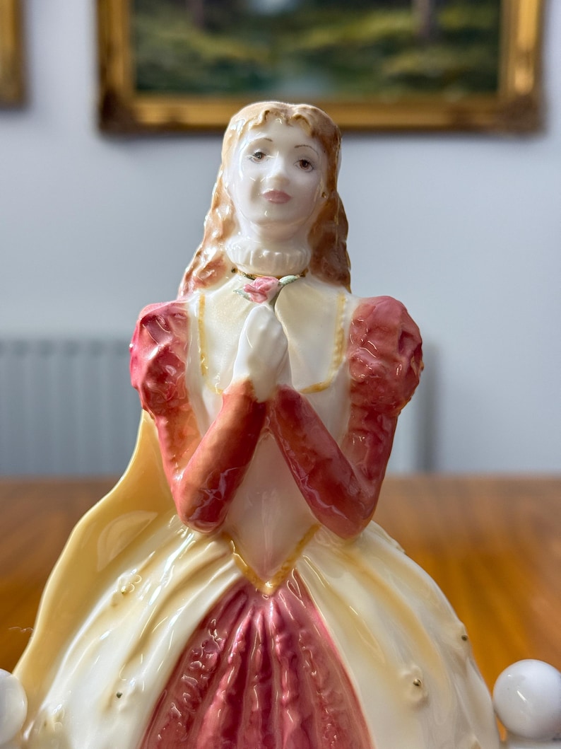 May include: A porcelain figurine of a woman in a long gown, with a cream-colored bodice and a pink skirt. The sleeves are a deep red, and she holds a pink rose. The figurine has long, flowing, light brown hair and is adorned with gold accents.