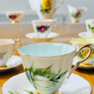 May include: A collection of vintage teacups and saucers with floral designs. The teacups have gold trim and handles, with a light blue interior. The saucers and teacups feature white roses with green leaves. A teapot and creamer are also visible.