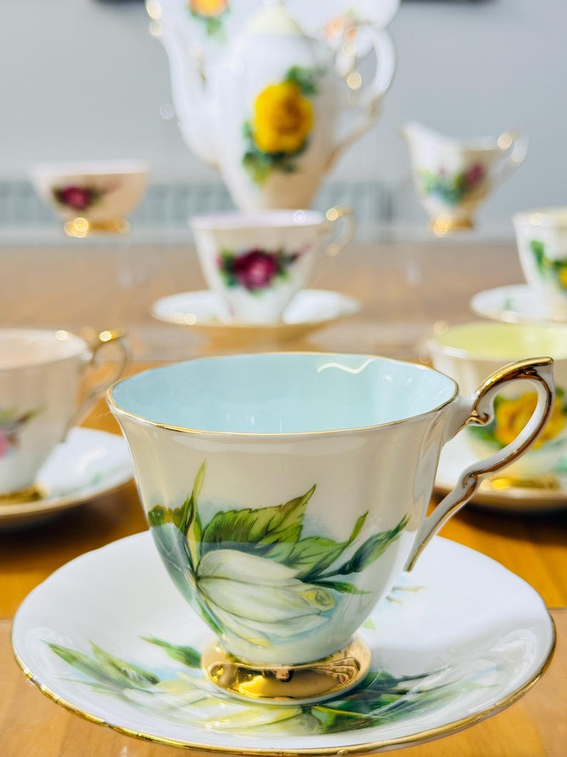 May include: A collection of vintage teacups and saucers with floral designs. The teacups have gold trim and handles, with a light blue interior. The saucers and teacups feature white roses with green leaves. A teapot and creamer are also visible.