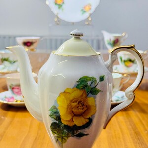 May include: White porcelain teapot with a yellow rose design. The teapot has a gold-colored base and handle accents. A matching plate and teacups with floral patterns are visible in the background, suggesting a tea set.