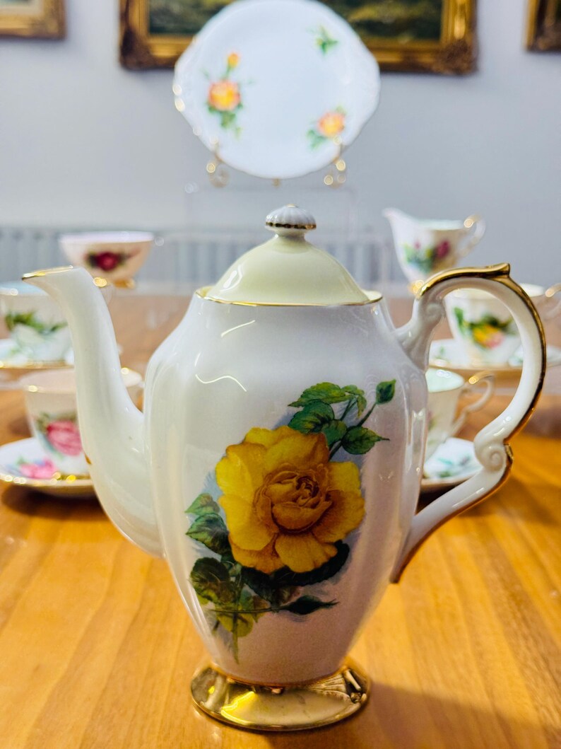 May include: White porcelain teapot with a yellow rose design. The teapot has a gold-colored base and handle accents. A matching plate and teacups with floral patterns are visible in the background, suggesting a tea set.