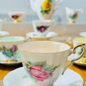 May include: A collection of vintage porcelain teacups and saucers, featuring floral designs with pink roses, green leaves, and gold trim. A teapot and creamer are also visible, creating a complete tea set.