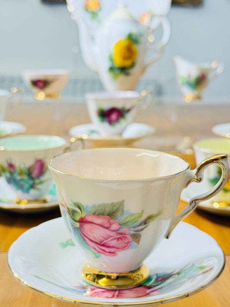 May include: A collection of vintage porcelain teacups and saucers, featuring floral designs with pink roses, green leaves, and gold trim. A teapot and creamer are also visible, creating a complete tea set.
