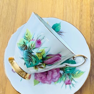 May include: White teacup and saucer set with a floral rose design in shades of pink, green, and teal. The teacup has a gold handle and trim, and the saucer has a scalloped edge. The set is likely vintage or antique.