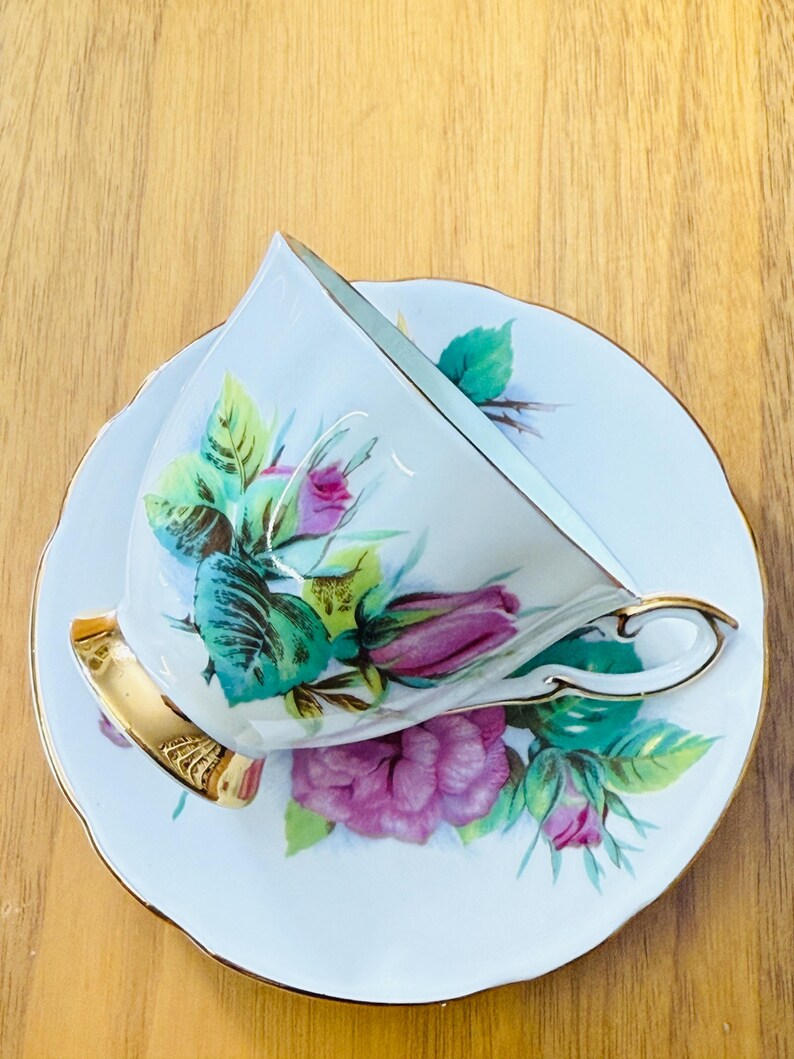 May include: White teacup and saucer set with a floral rose design in shades of pink, green, and teal. The teacup has a gold handle and trim, and the saucer has a scalloped edge. The set is likely vintage or antique.