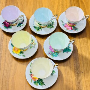 May include: Six vintage teacups and saucers arranged on a wooden surface. Each cup is a different pastel color: lavender, light blue, peach, yellow, mint green, and pale yellow. The saucers feature floral designs with gold trim.