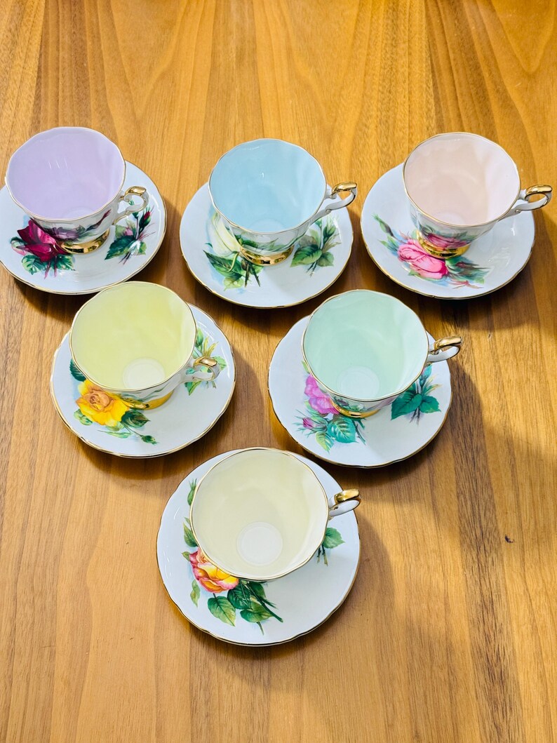 May include: Six vintage teacups and saucers arranged on a wooden surface. Each cup is a different pastel color: lavender, light blue, peach, yellow, mint green, and pale yellow. The saucers feature floral designs with gold trim.