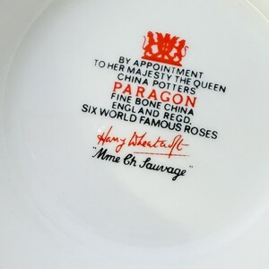 May include: Close-up of a white Paragon fine bone china piece with red text and a crest. The text includes "By Appointment to Her Majesty The Queen" and "Six World Famous Roses". The signature "Hany Wheatcroft" is also visible.