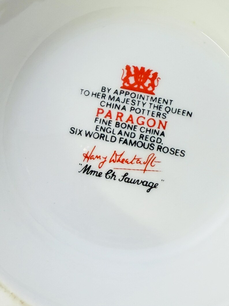 May include: Close-up of a white Paragon fine bone china piece with red text and a crest. The text includes "By Appointment to Her Majesty The Queen" and "Six World Famous Roses". The signature "Hany Wheatcroft" is also visible.